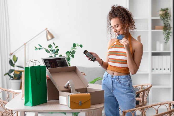 Young woman with credit card shopping online on her phone, surrounded by boxes and bags of goods.